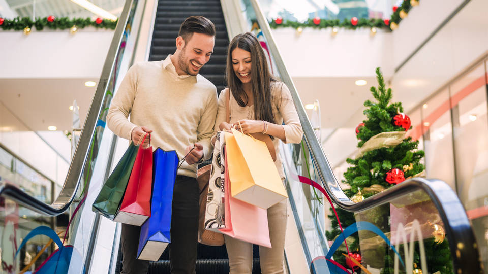 male female millennial couple descend holiday decorated retail mall escalator carrying purchases in shopping bags