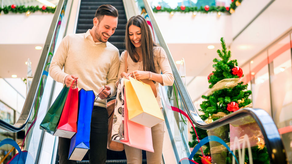 male and female couple shoppers carying multiple shopping bags descending escalator in retail shopping mall decorated for winter holidays