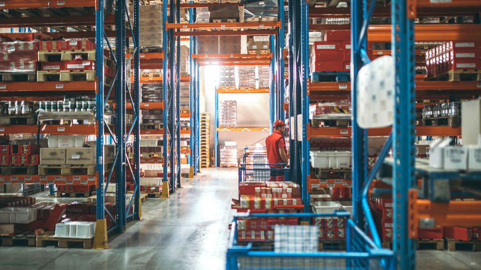 worker in hard had and safety vest in large warehouse with multilevel shelving in orange and blue steel