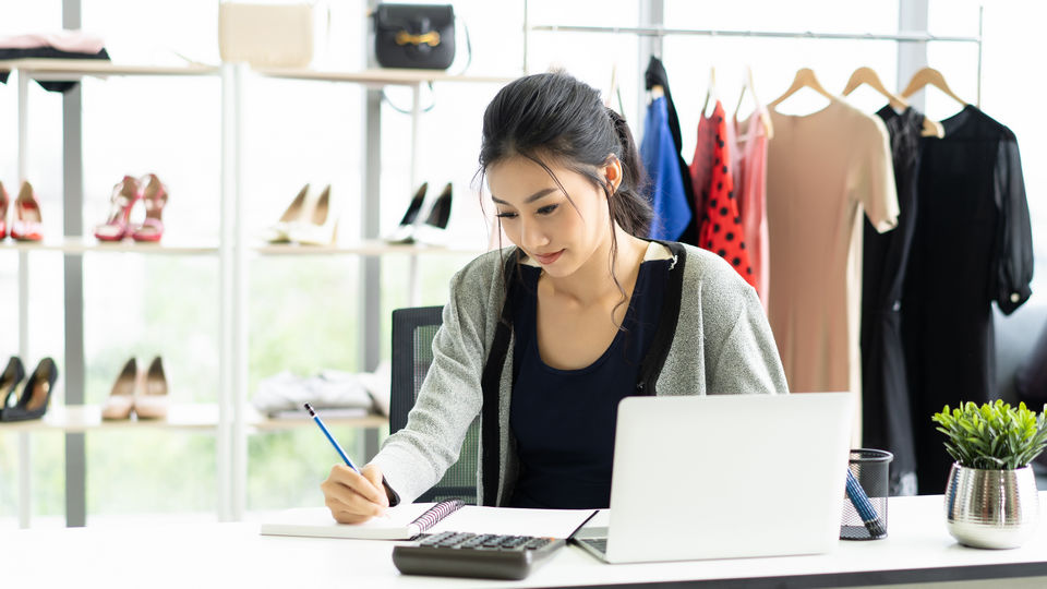 female retail store manager reviewing store data on laptop while writing with pen on paper