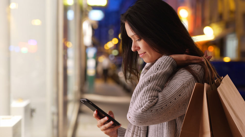 woman carrying shopping bags looking at cell phone in front of retail store window on shopping street in the evening