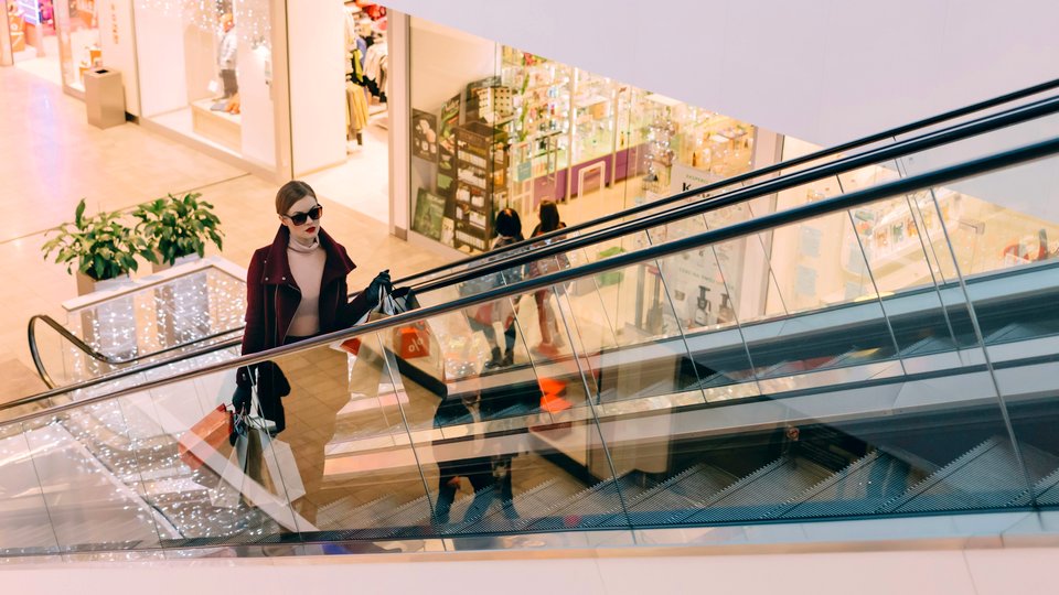 female shopper wearing sunglasses and carrying several shopping bags on the up escalator in a retail shopping mall