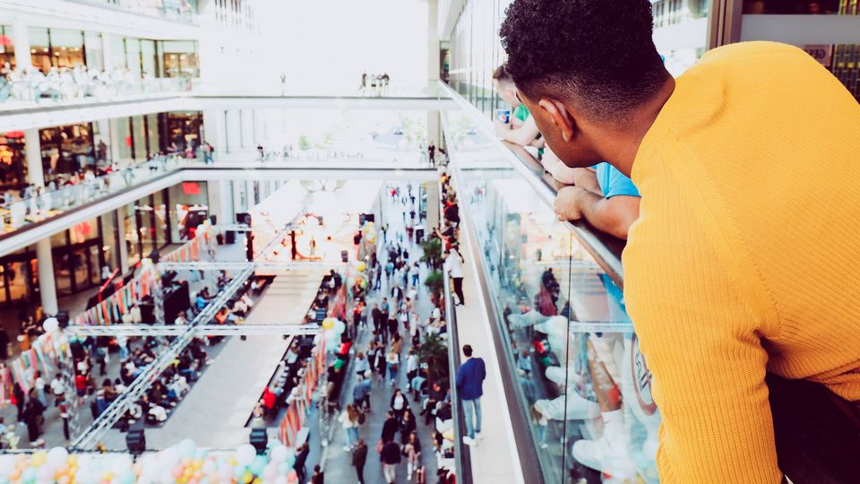 man shopping in multi-level retail mall looking for friend leans over the open atrium to the ground level