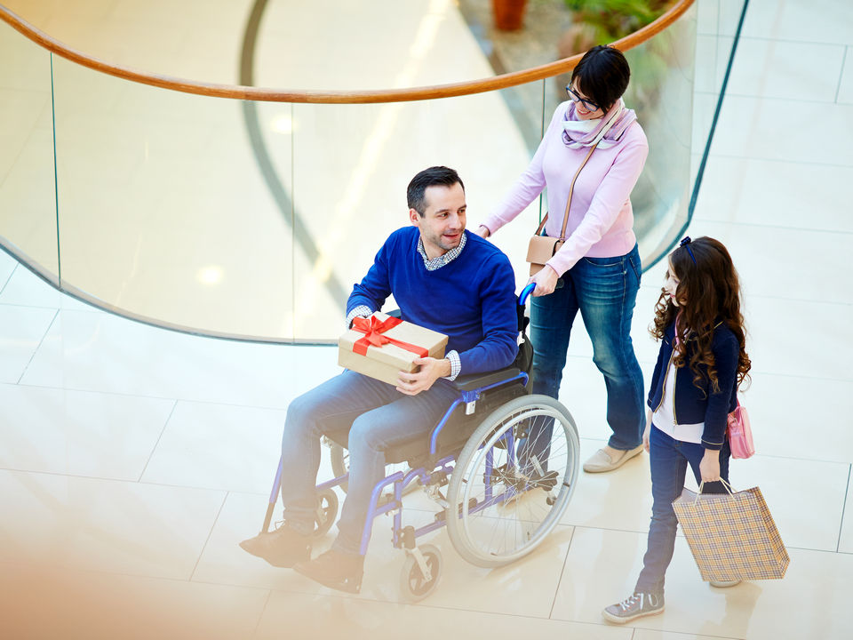 disabled man in wheelchair holds a gift box purchase while his wife and daughter accompany him through retail shopping mall