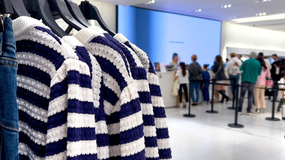 busy retail apparel store with sweaters on a rack in the foreground and several queues of shopper at register in background