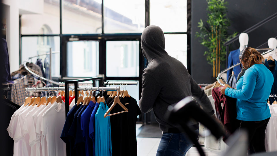 shoplifter in hoodie stealing merchandise from retail apparel store while other shoppers do not notice